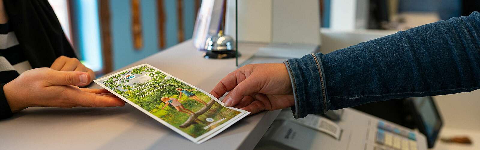 <p>Mitarbeiter der Touristinfo übergibt Infobroschüre an Kunde</p>
